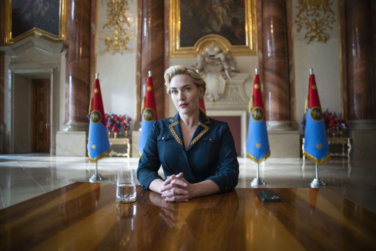 A woman in a blue dress sits at a table with flags behind her.