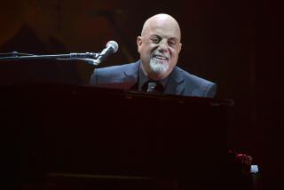 Billy Joel in a suit sitting at a piano on a stage smiling with a microphone in front of his face