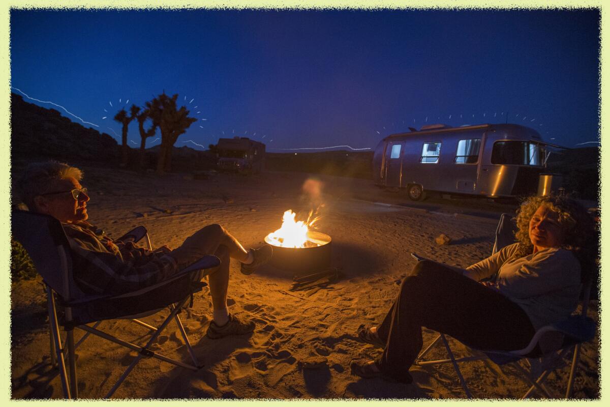 Two people sit next to a campfire in a desert campground area; a trailer sits in the background. 