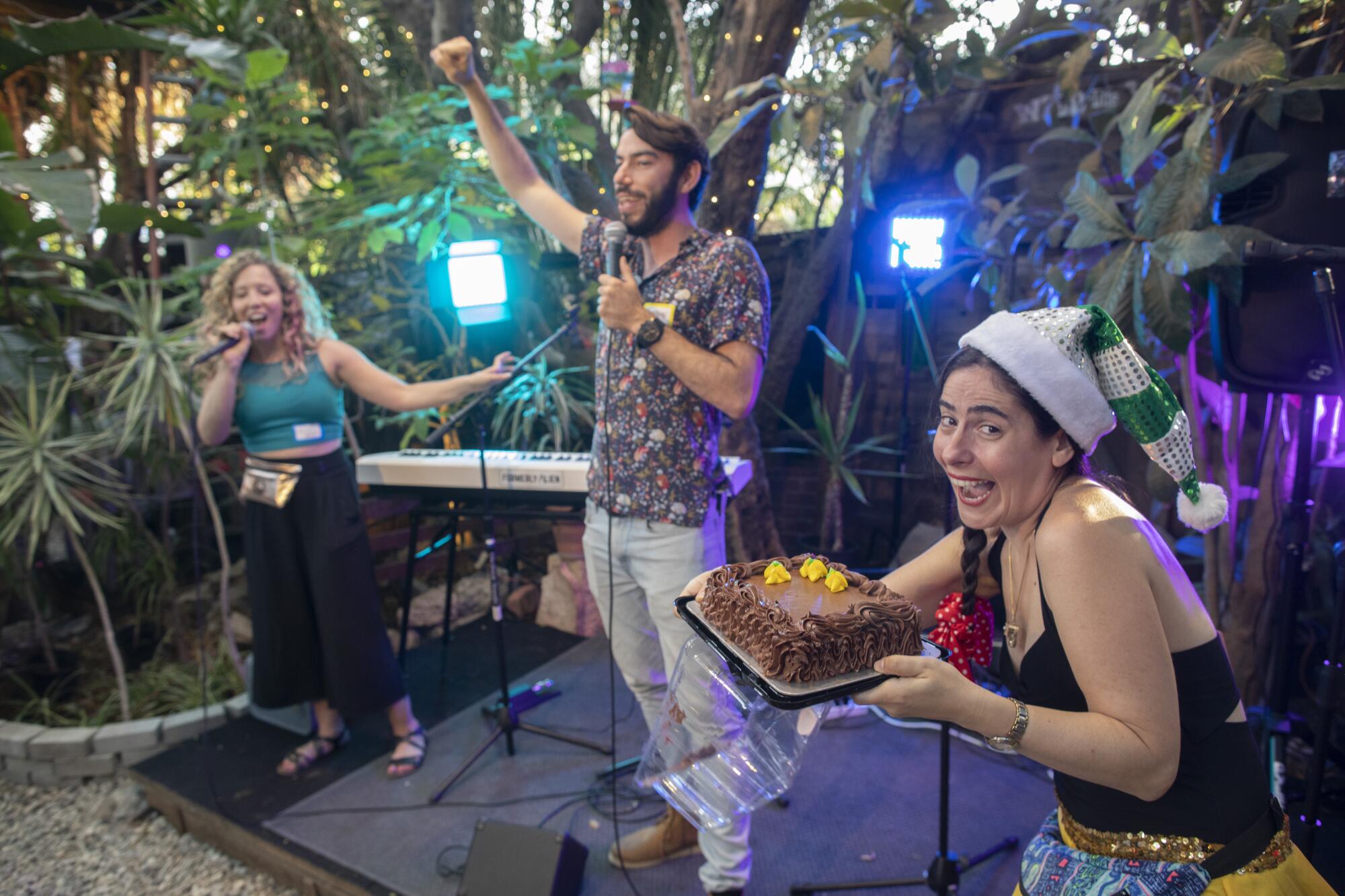 A woman, right, carries a cake as a man, center, and another woman gesture while holding microphones 
