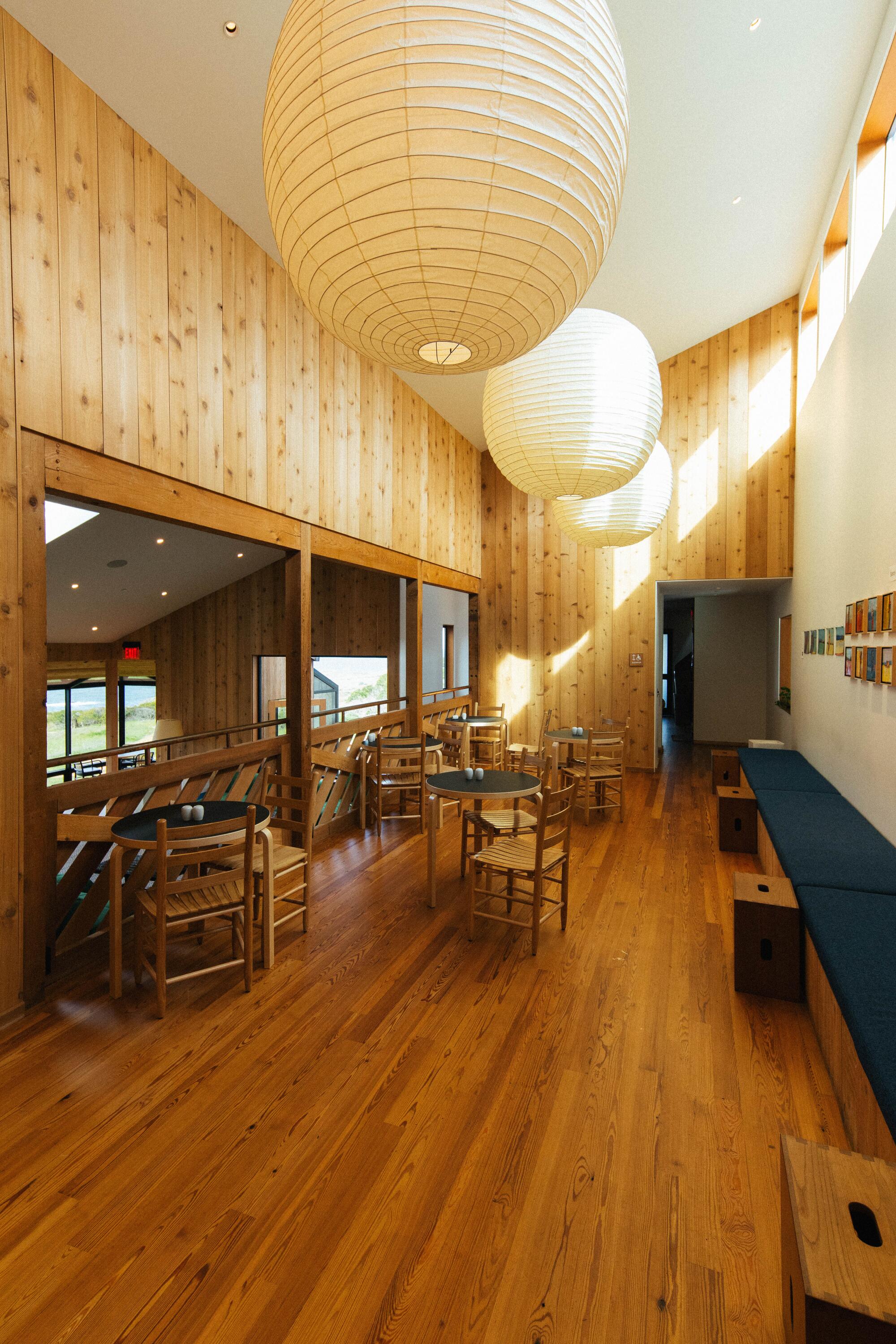 A room with wood walls and floor, tables and chairs, and large hanging paper light fixtures.