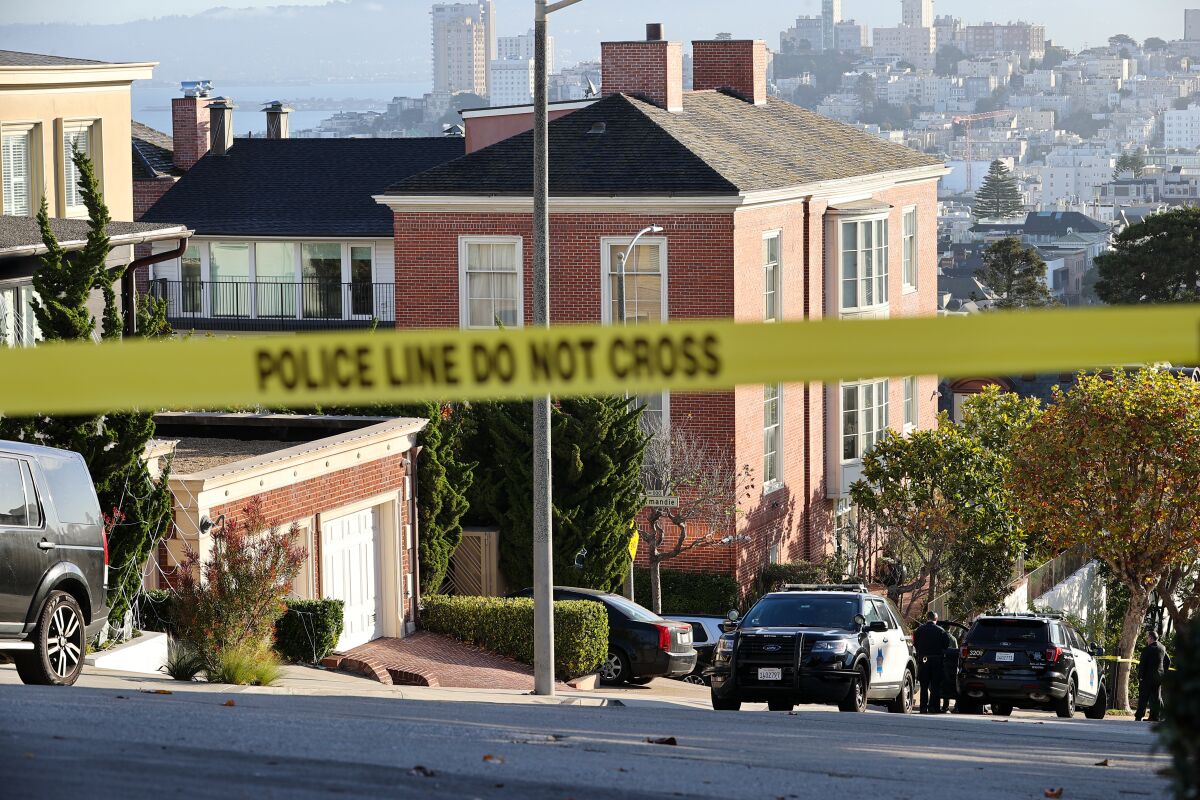 Police tape is seen in the foreground. In back, police units are parked alongside a stately mansion on a city street.