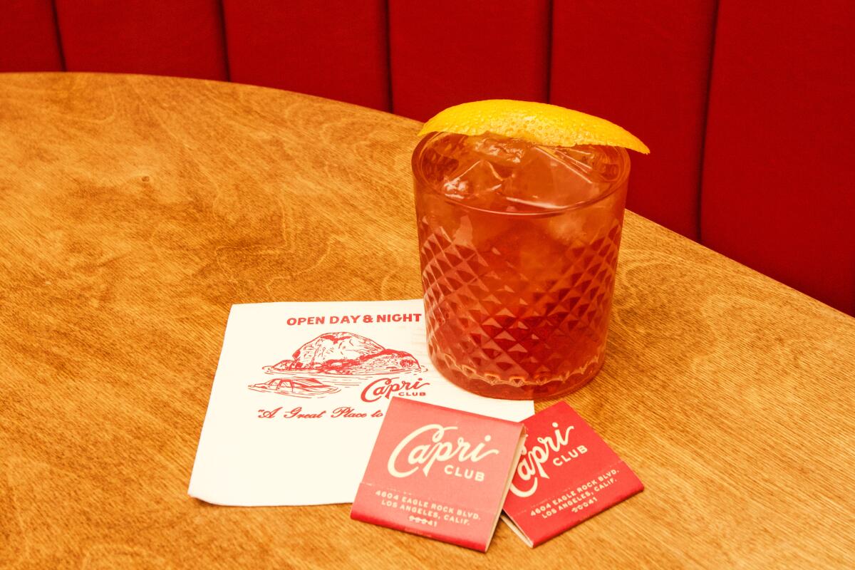 A photo of a negroni in a crystal tumbler, garnished by orange slice, sitting next to branded matchbooks and napkin.