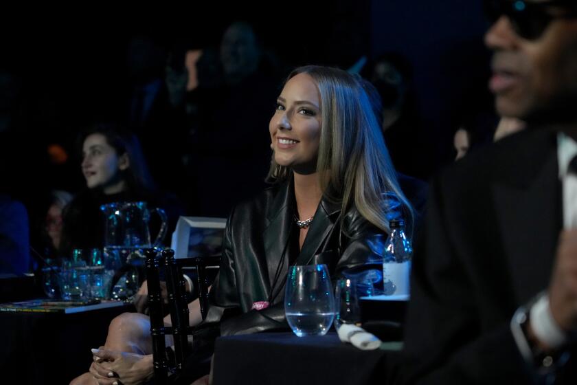 A smiling young woman with long hair sits amid other people at a table in a darkened room