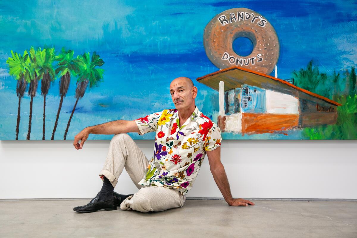 A man wearing a floral shirt sits on the floor before a painting of palm trees and Randy's Donuts.