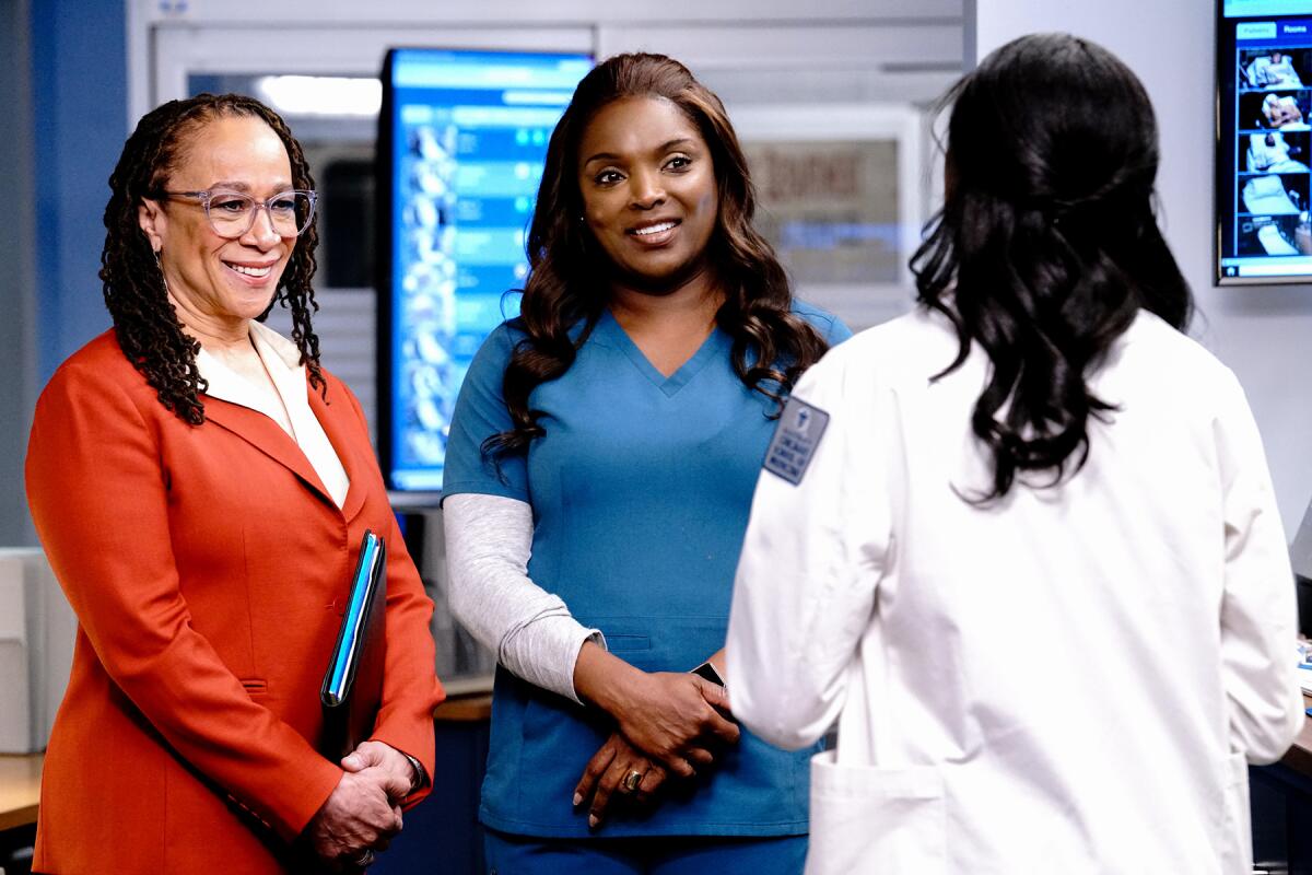 Two women, one in a business suit and another in scrubs, talk to another woman in a white coat with her back turned.