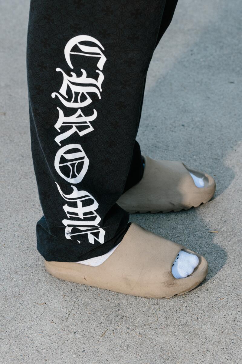 A closeup of a pair of legs wearing black Chrome Hearts sweats, tan slides and white socks.