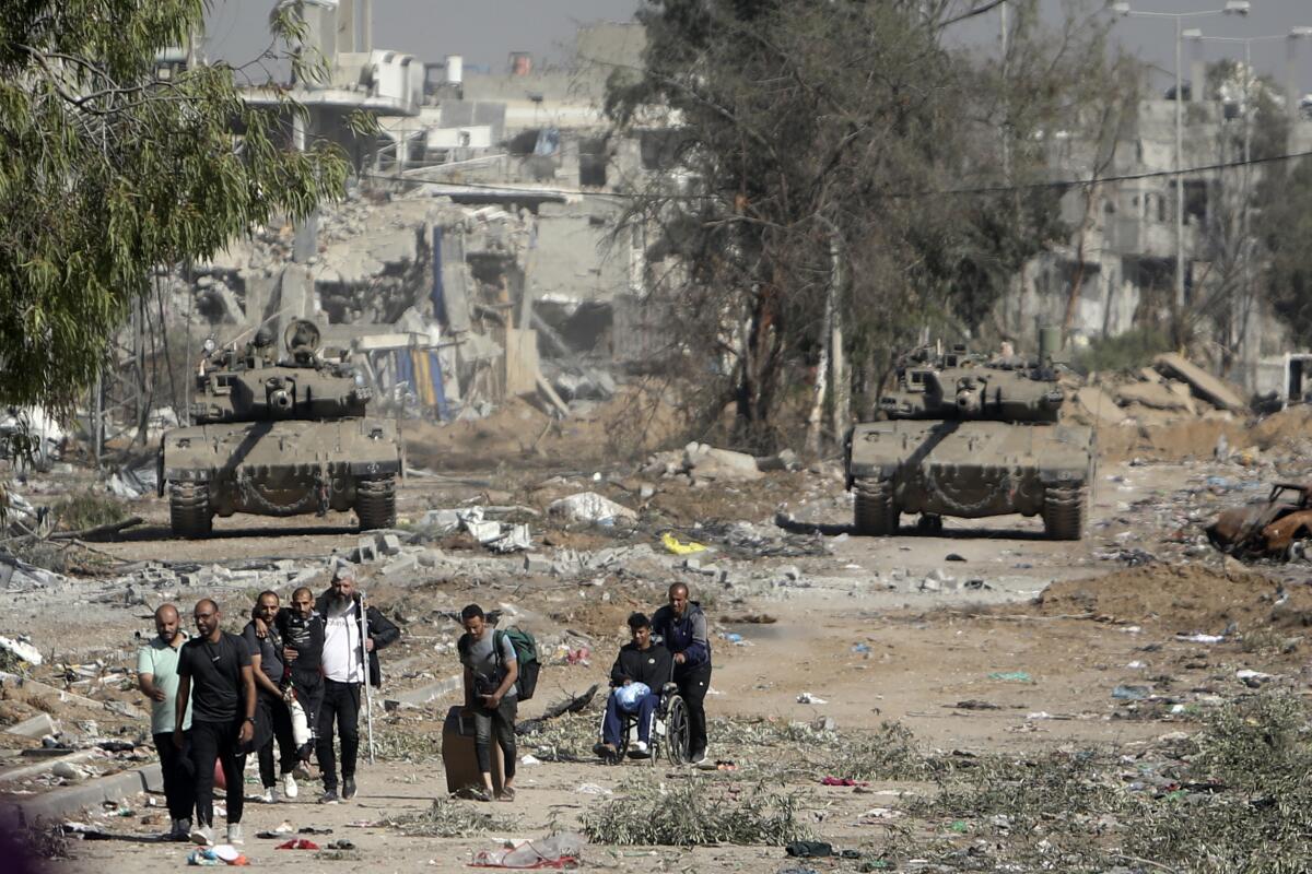 Palestinians walk over dirt with rubble and two tanks in the background.
