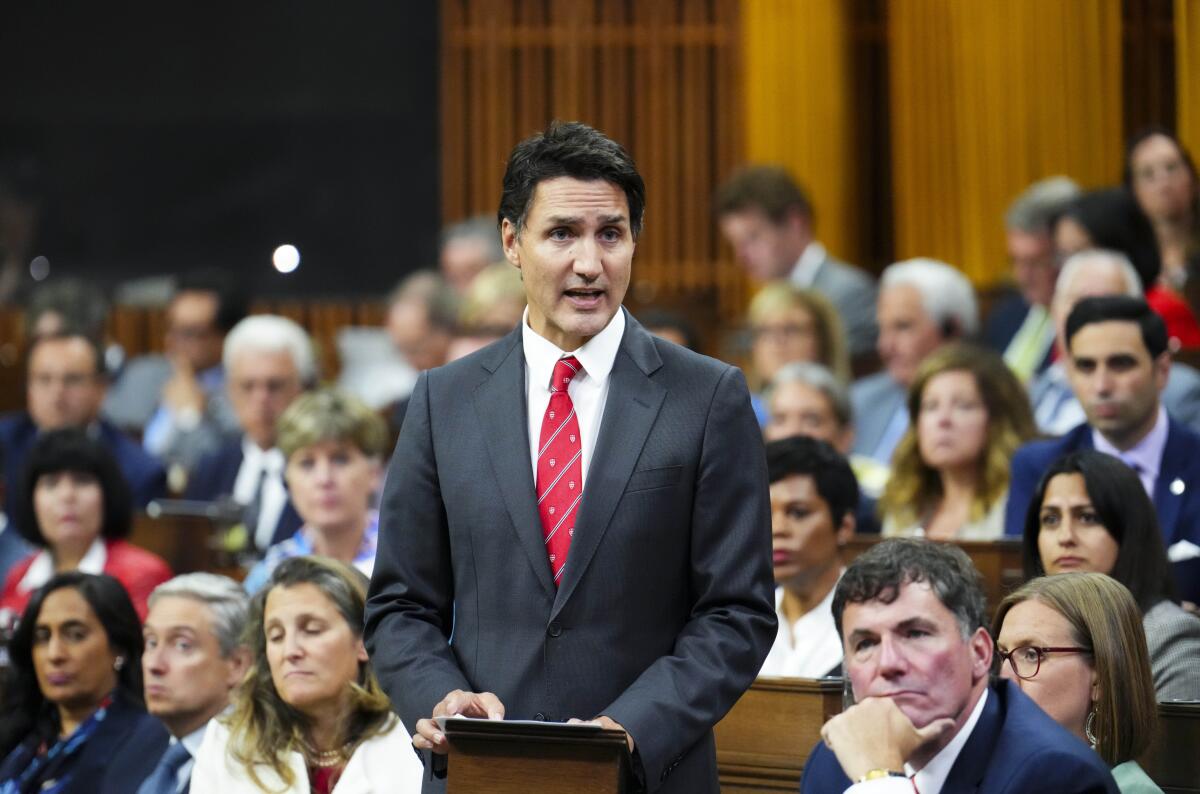 A man with dark hair, in a dark gray suit and red tie, speaks while standing amid a seated crowd