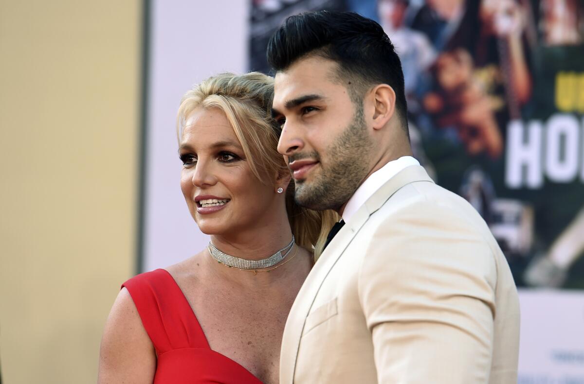 Britney Spears and Sam Asghari stand tightly together and look off to the left 