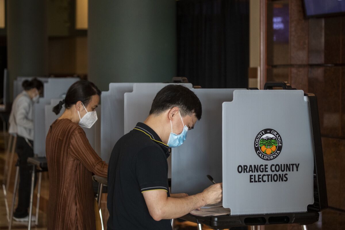 Three people vote on election day, wearing masks. The voting booths bear the seal of Orange County, California.