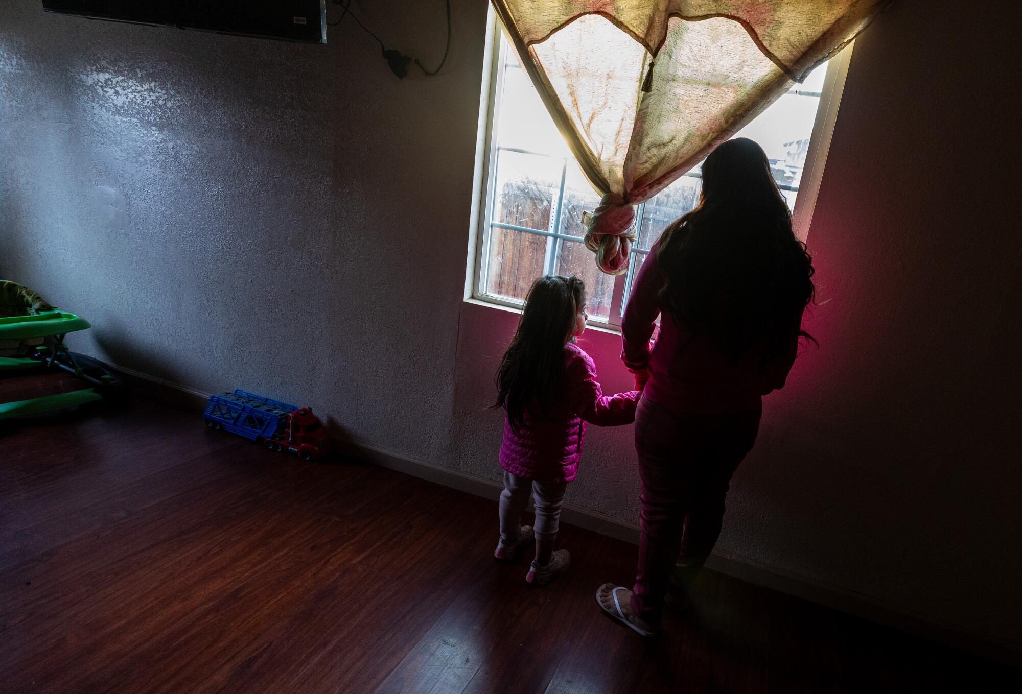 Woman and child look out the window of a sparsely furnished house.