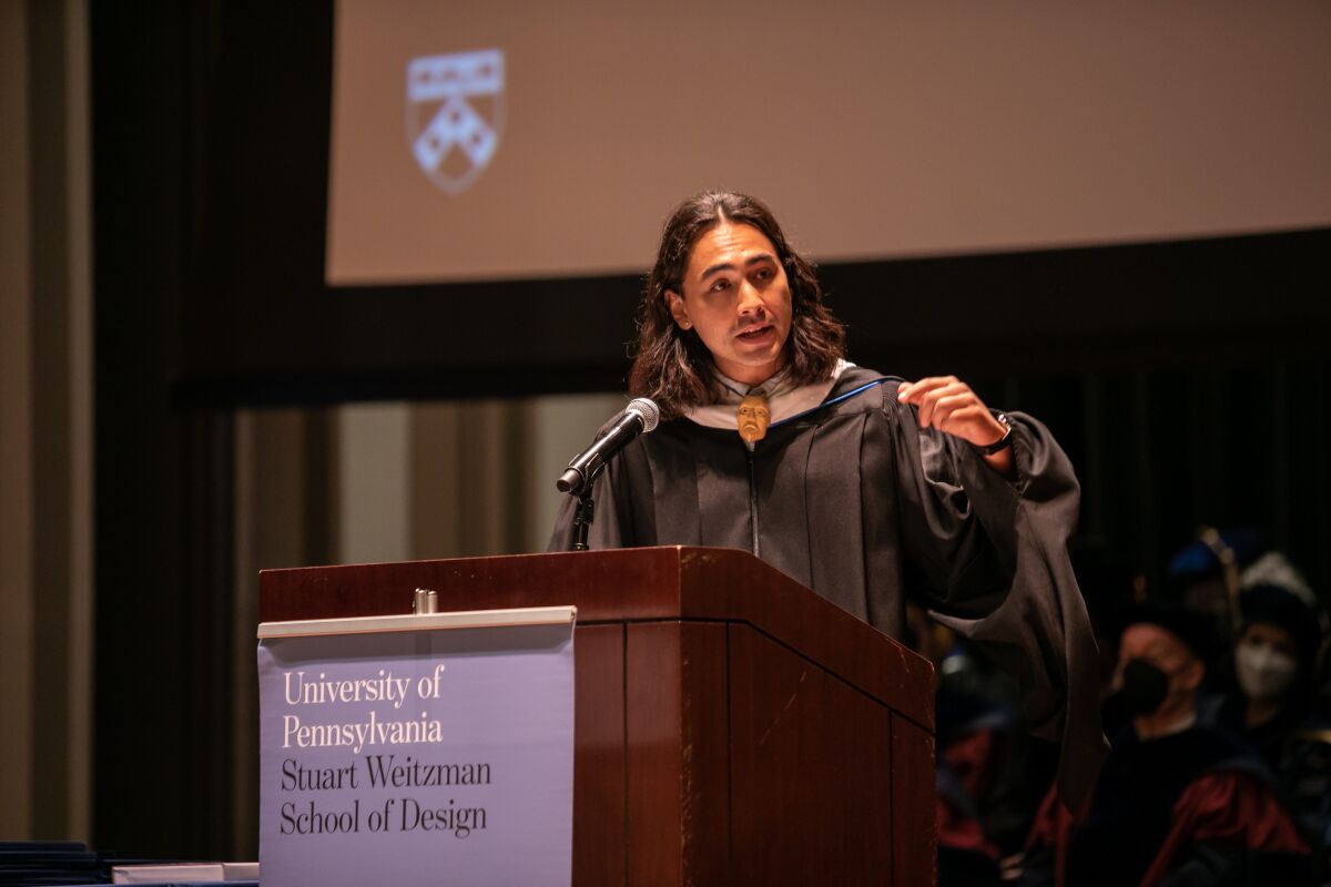 A man in a graduation gown speaks at a podium.