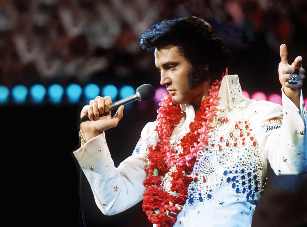 Elvis Presley performs wearing a red lei and a white jumpsuit with a colorful bejeweled design.