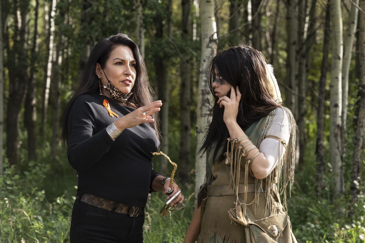 A woman confers with "Prey" star Amber Midthunder dressed in 1719 Comanche clothing in the forest setting of the film.
