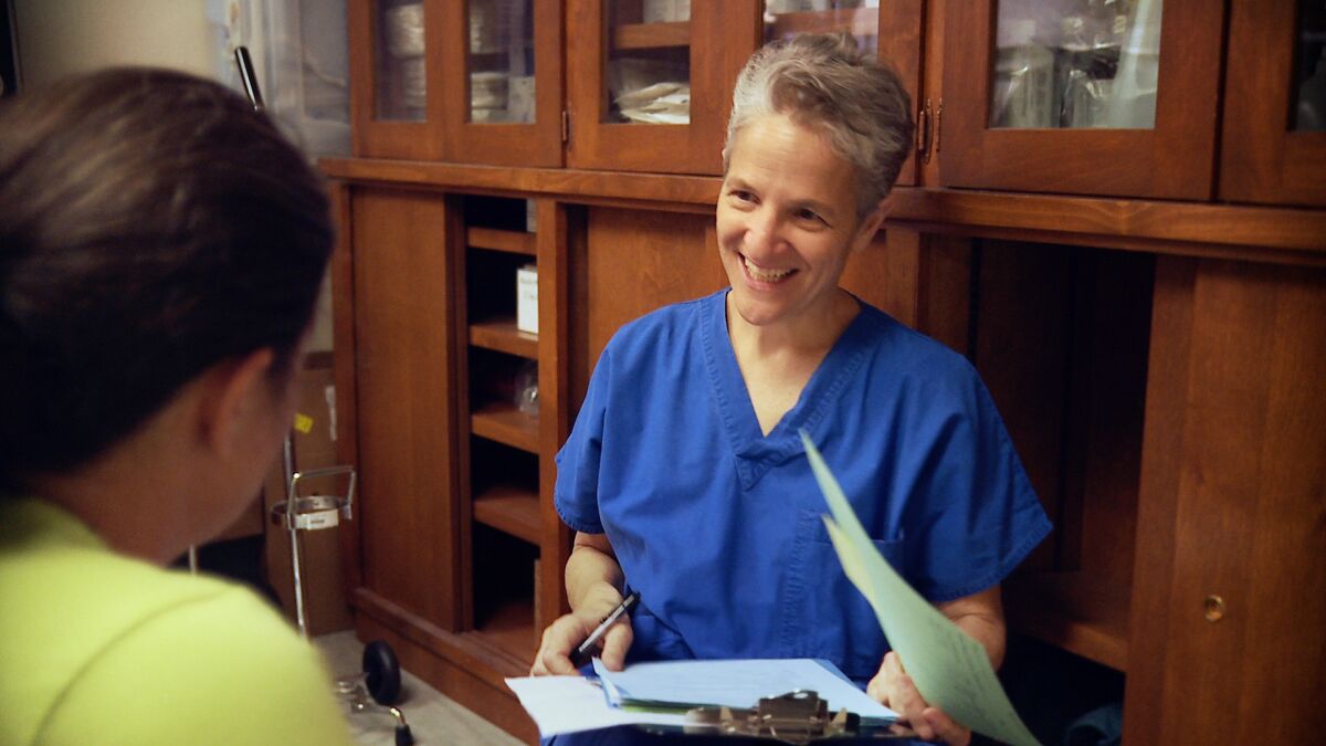 A doctor in blue scrubs holding a clipboard while meeting with a patient 