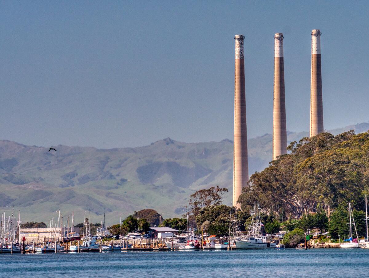 Three tall smoke stacks tower over the landscape, with a harbor in the foreground.