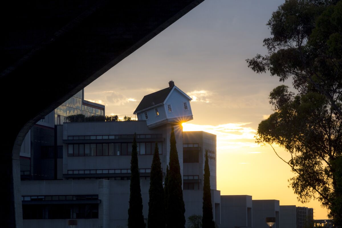 A pale blue house sits at a tilt on the edge of a massive concrete campus building in the dusk light.