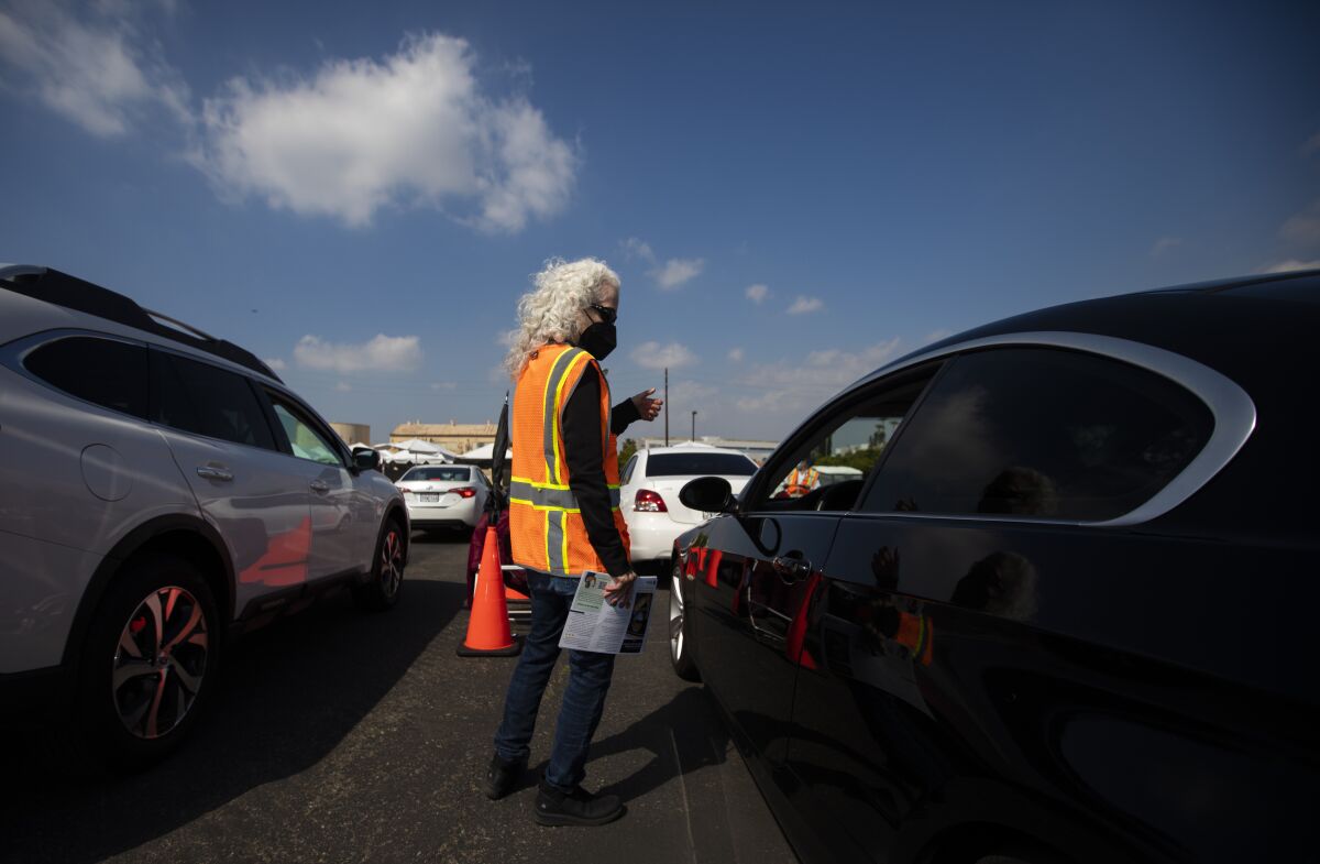 Barbara Ferrer in a black mask and orange vest stands between cars and talks to passengers.