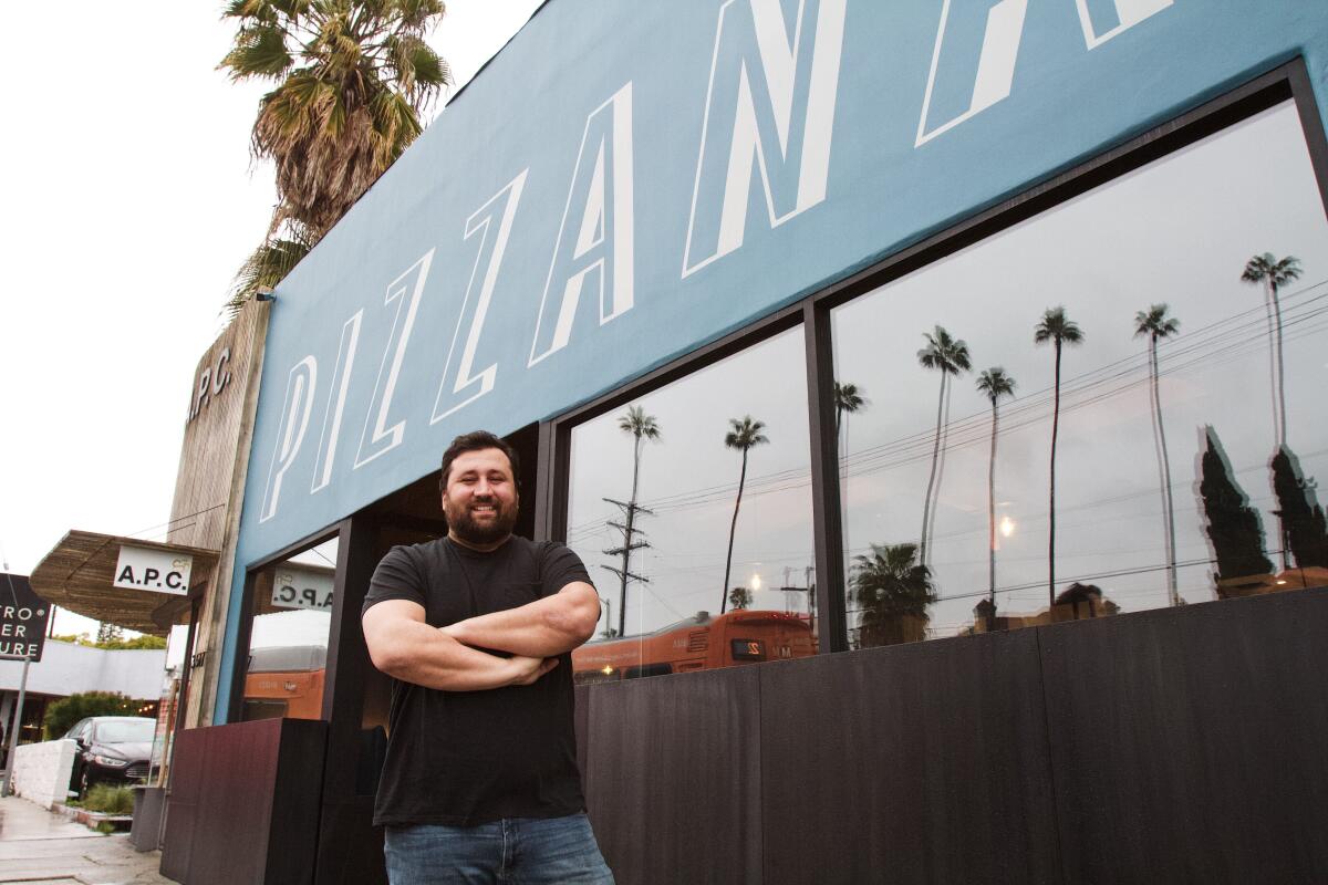 A man in a black shirt stands outside a black-and-blue restaurant that says "Pizzana." Palm trees reflect in the windows
