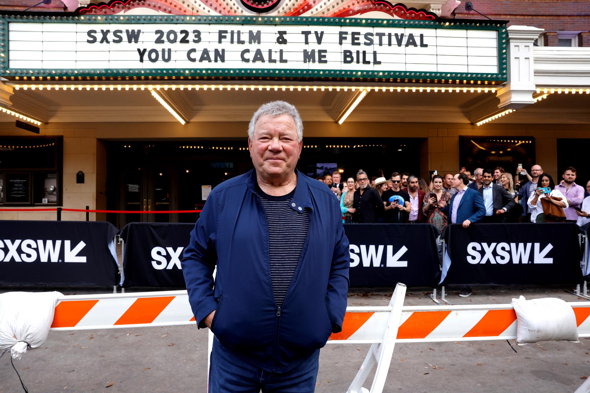 William Shatner stands in front of a movie theater marquee in a blue jacket and black T-shirt.
