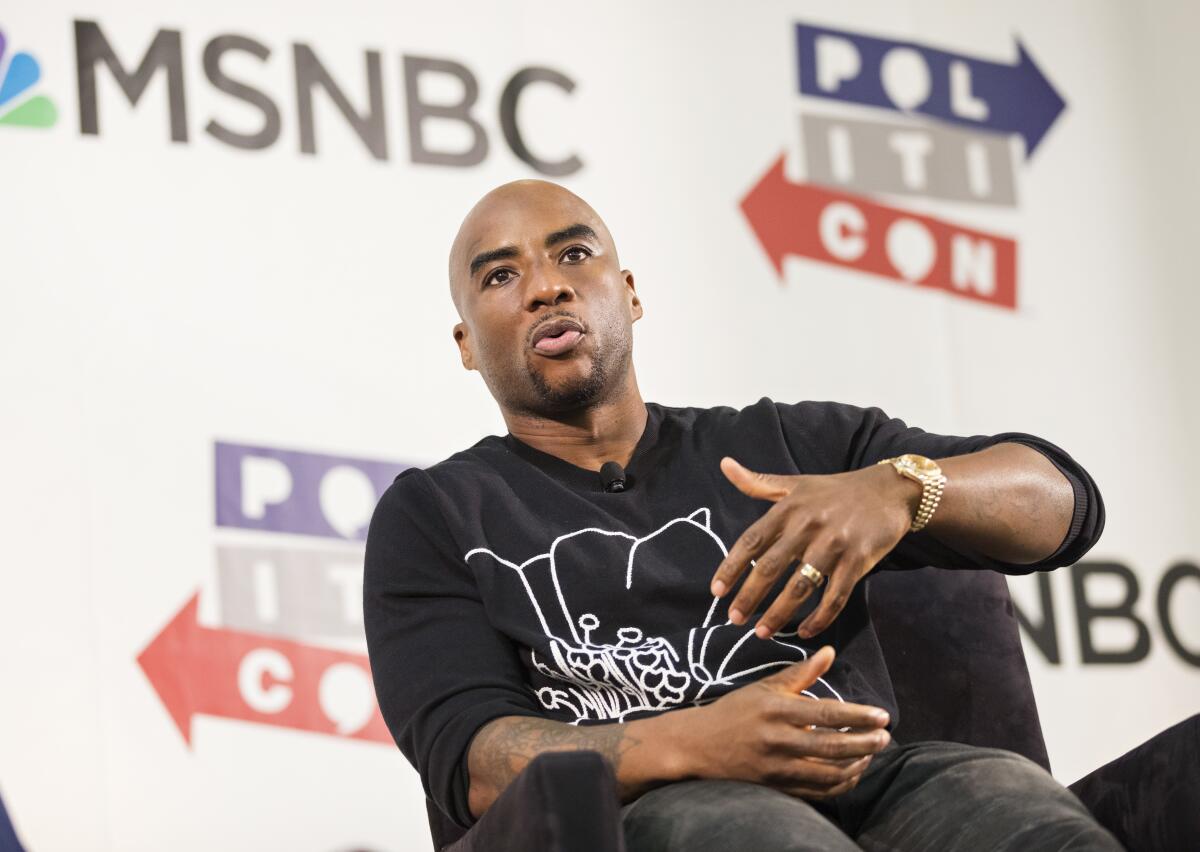 Charlamagne Tha God sits and speaks in a chair onstage in a black shirt with his hands in front of him