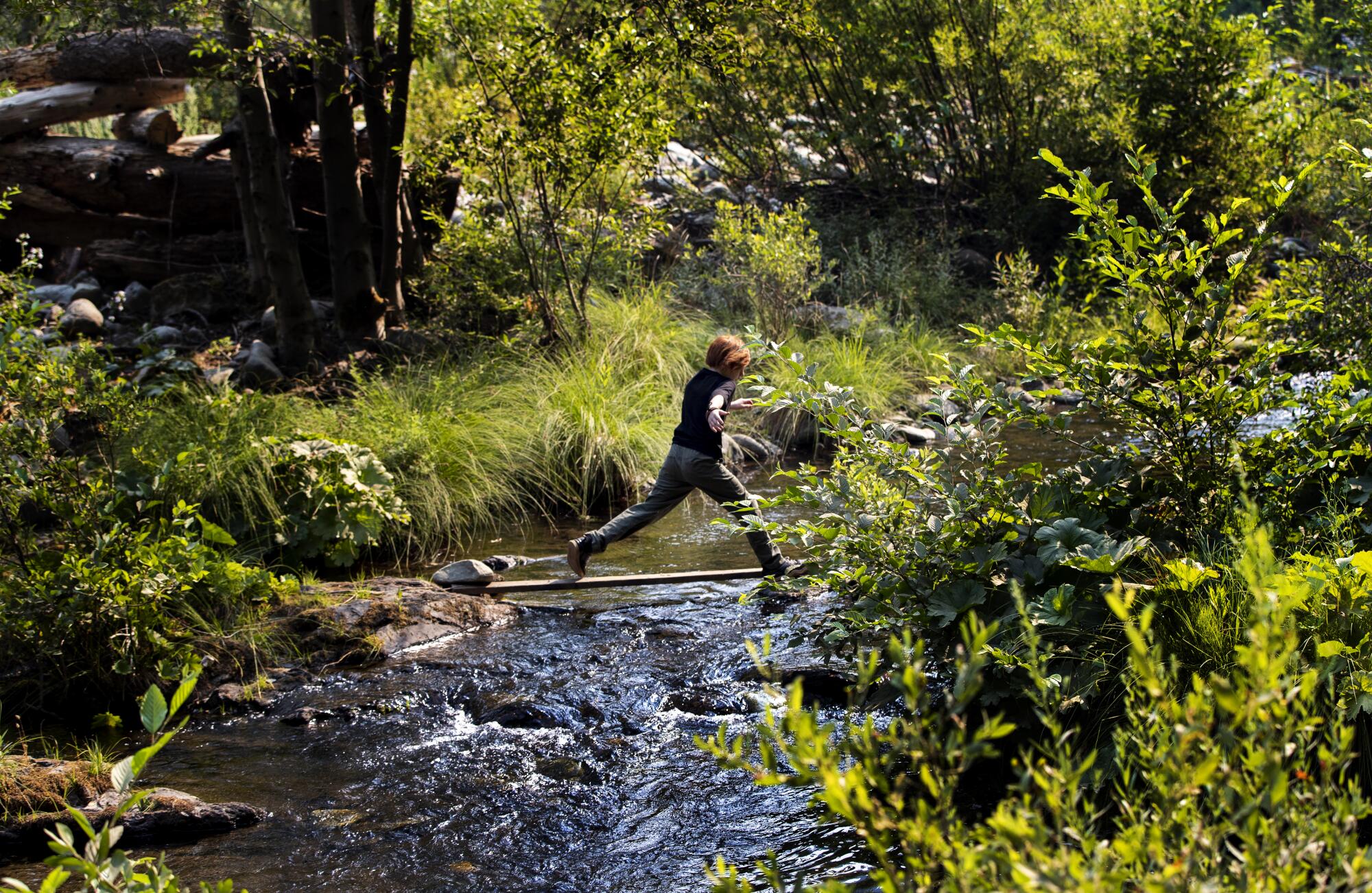 A boy leaps across a quickly moving stream bordered by green banks. 