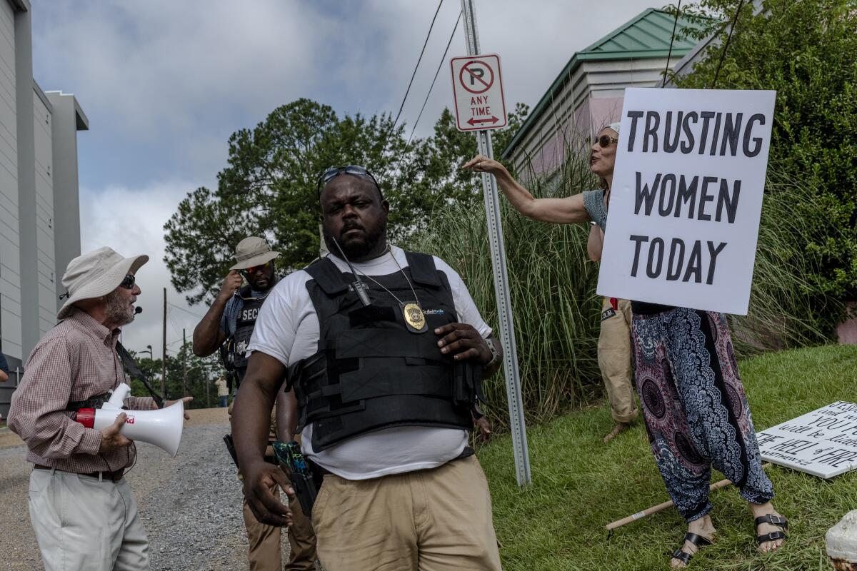 A woman holds a sign that reads Trusting Women Today across from a man with a bullhorn. Security officers stand between them.