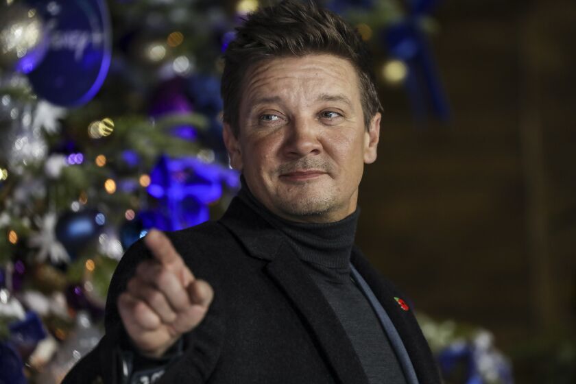 Jeremy Renner in a black blazer and dark turtleneck smiling and pointing his right hand