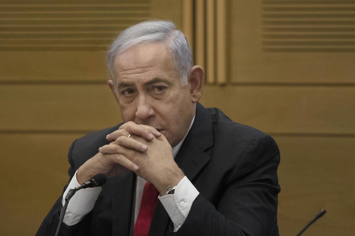 Benjamin Netanyahu sits at a microphone with folded hands.