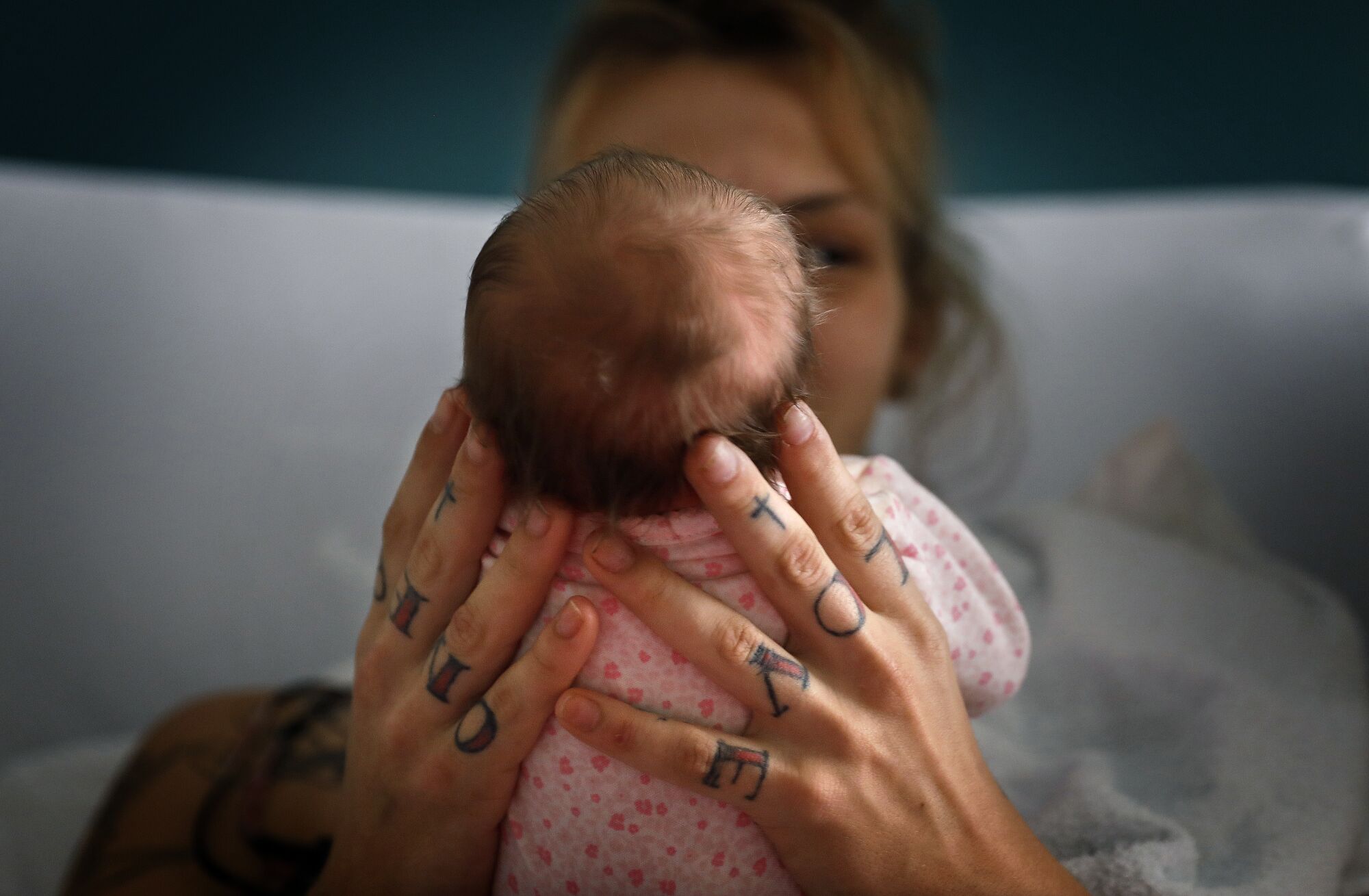 A woman with tattoos on her knuckles holding a baby as seen from behind the baby's head.