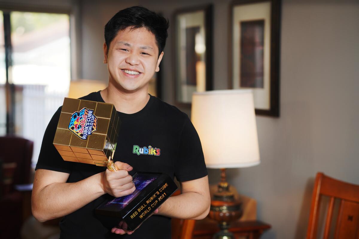 A young man poses for the camera, smiling, while holding a trophy modeled after a Rubik's Cube.
