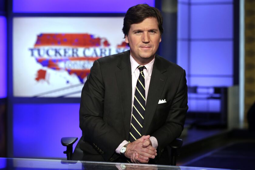 Tucker Carlson wears a suit and folds his hands on a desk