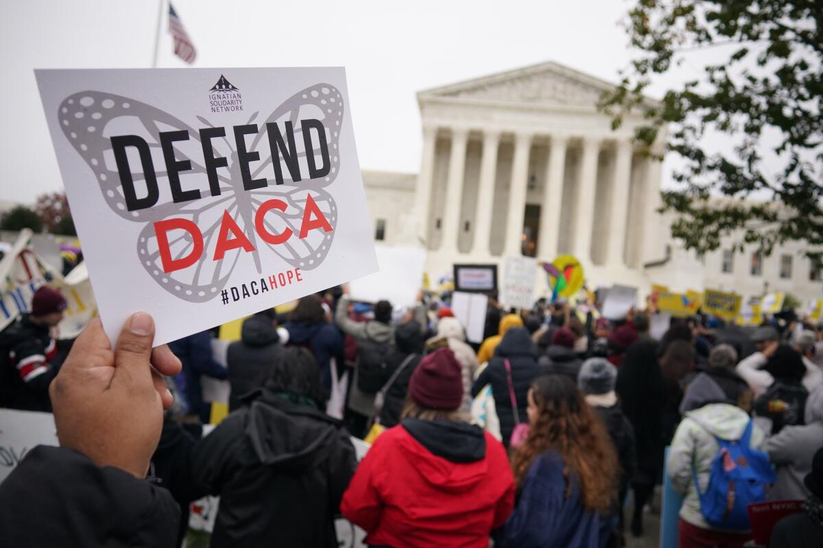 A hand holds a sign reading "Defend DACA / #DACAHOPE" over a butterfly design as a crowd protests outside the Supreme Court.