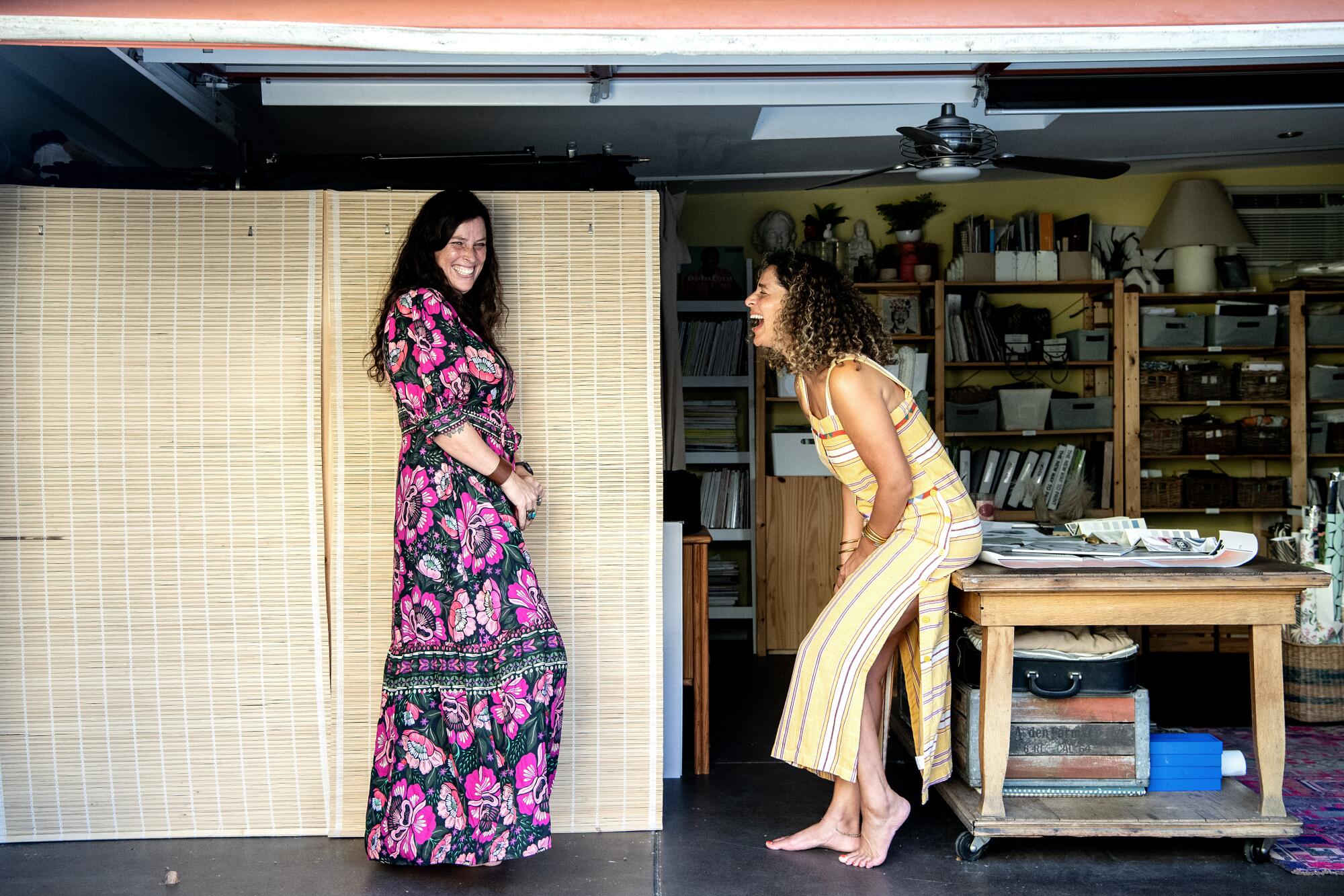 Two women stand in a garage-turned-workspace, laughing.