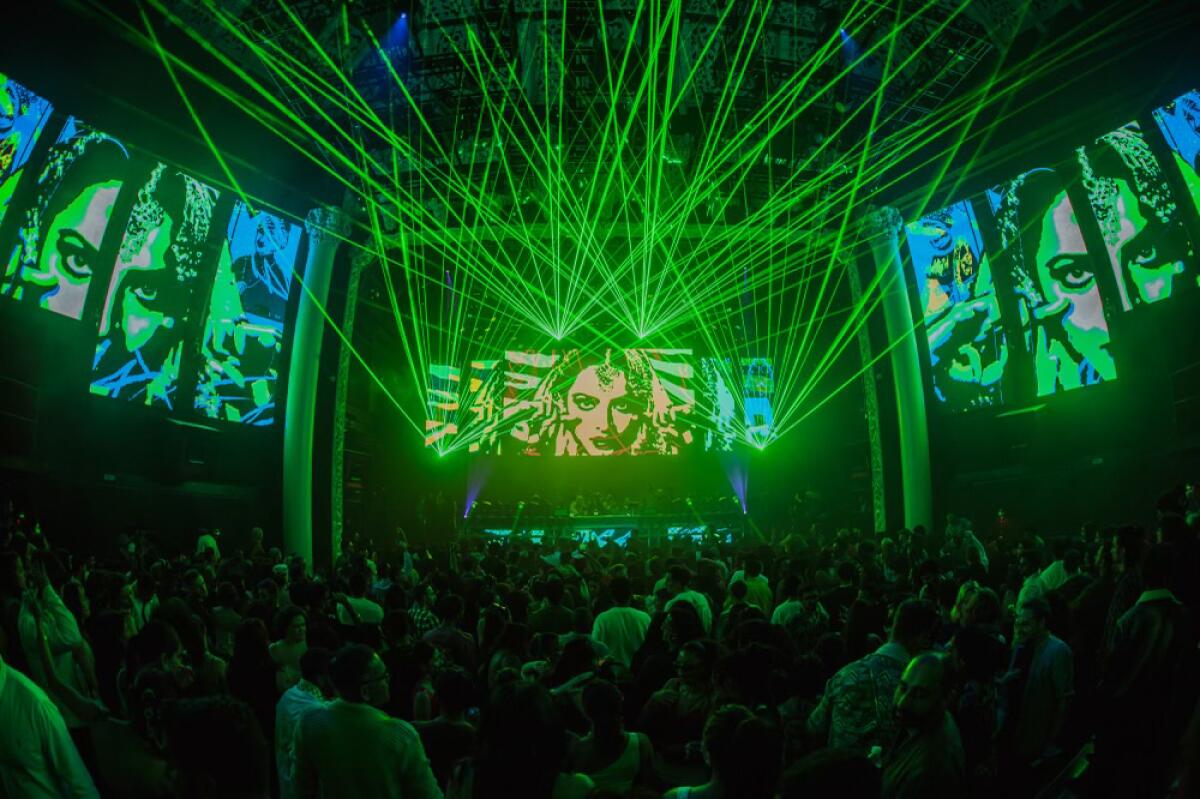 A large crowd jamming out under green lights and video effects as a DJ plays music.