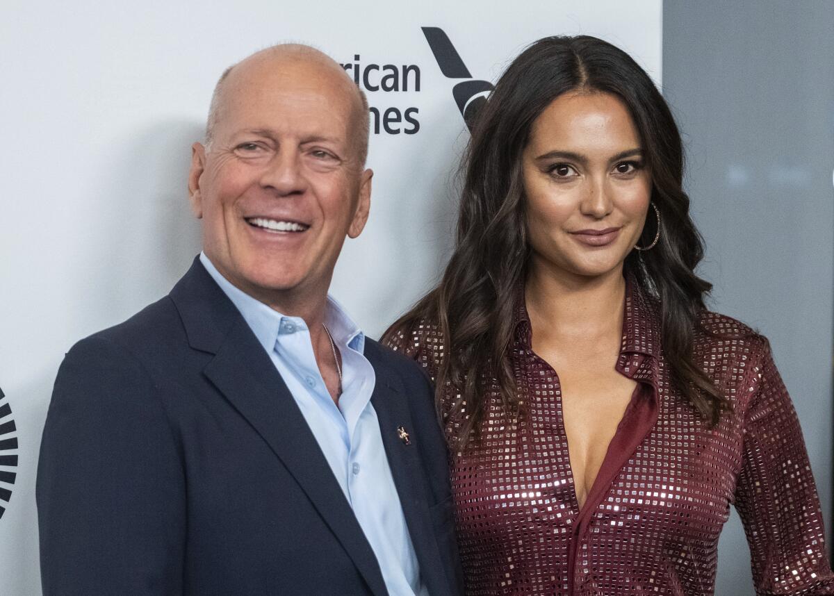 Bruce Willis in a dark suit jacket and blue suit shirt smiling next to a woman with dark brown hair in a maroon dress