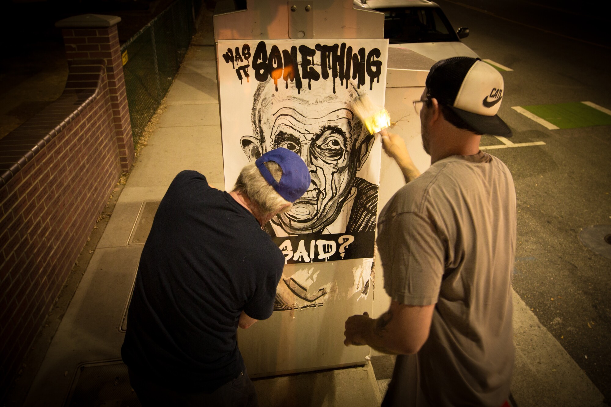 Robbie Conal and a man in a baseball cap paste a poster of Rudolph Giuliani onto an electrical box at night.