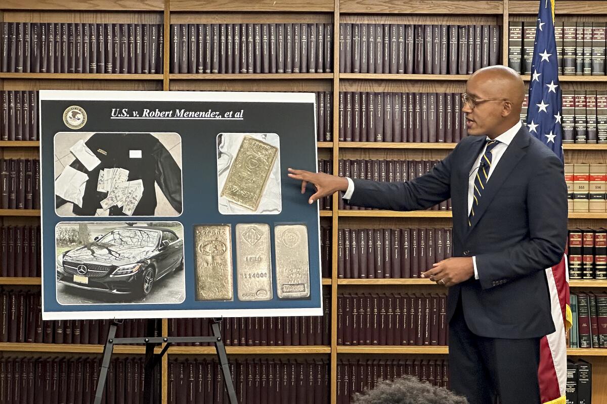 A man in a suit points to a poster board with photos and the title, "U.S. v. Robert Menendez, et al"