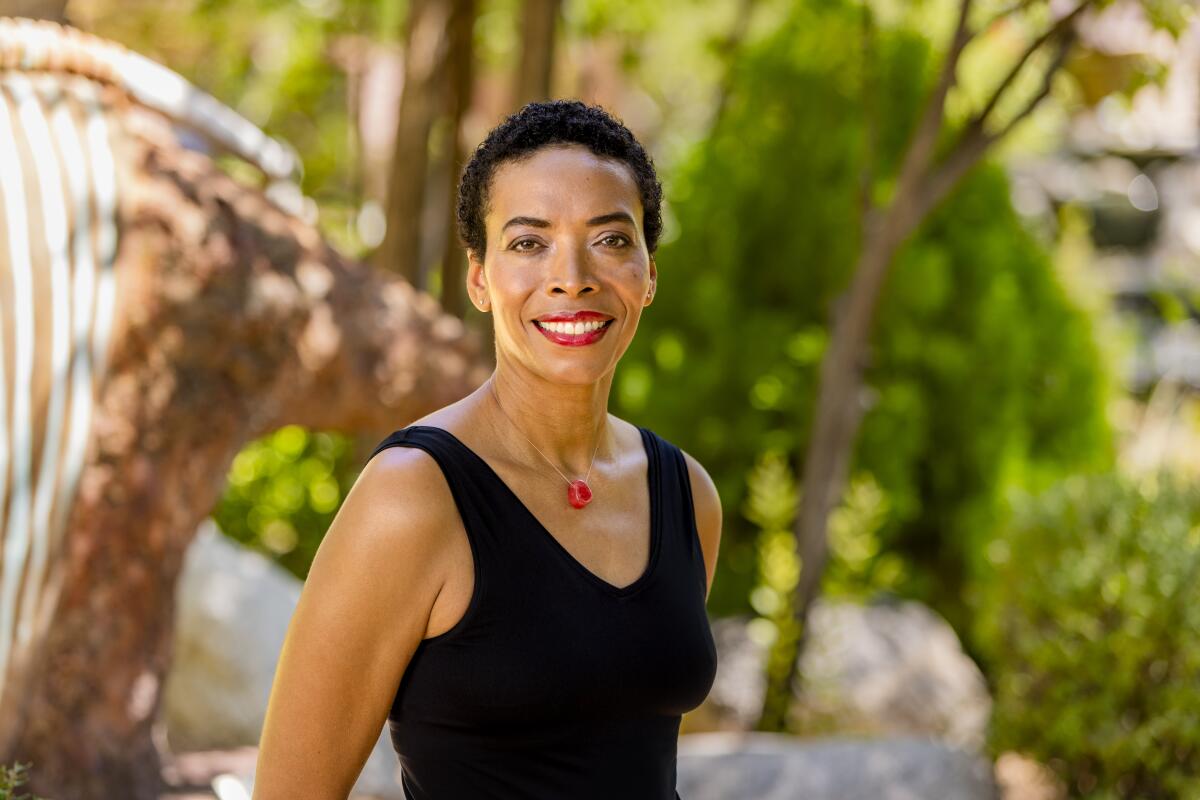 A brown-skinned woman in a black tank top with close-cropped hair smiling in front of trees.
