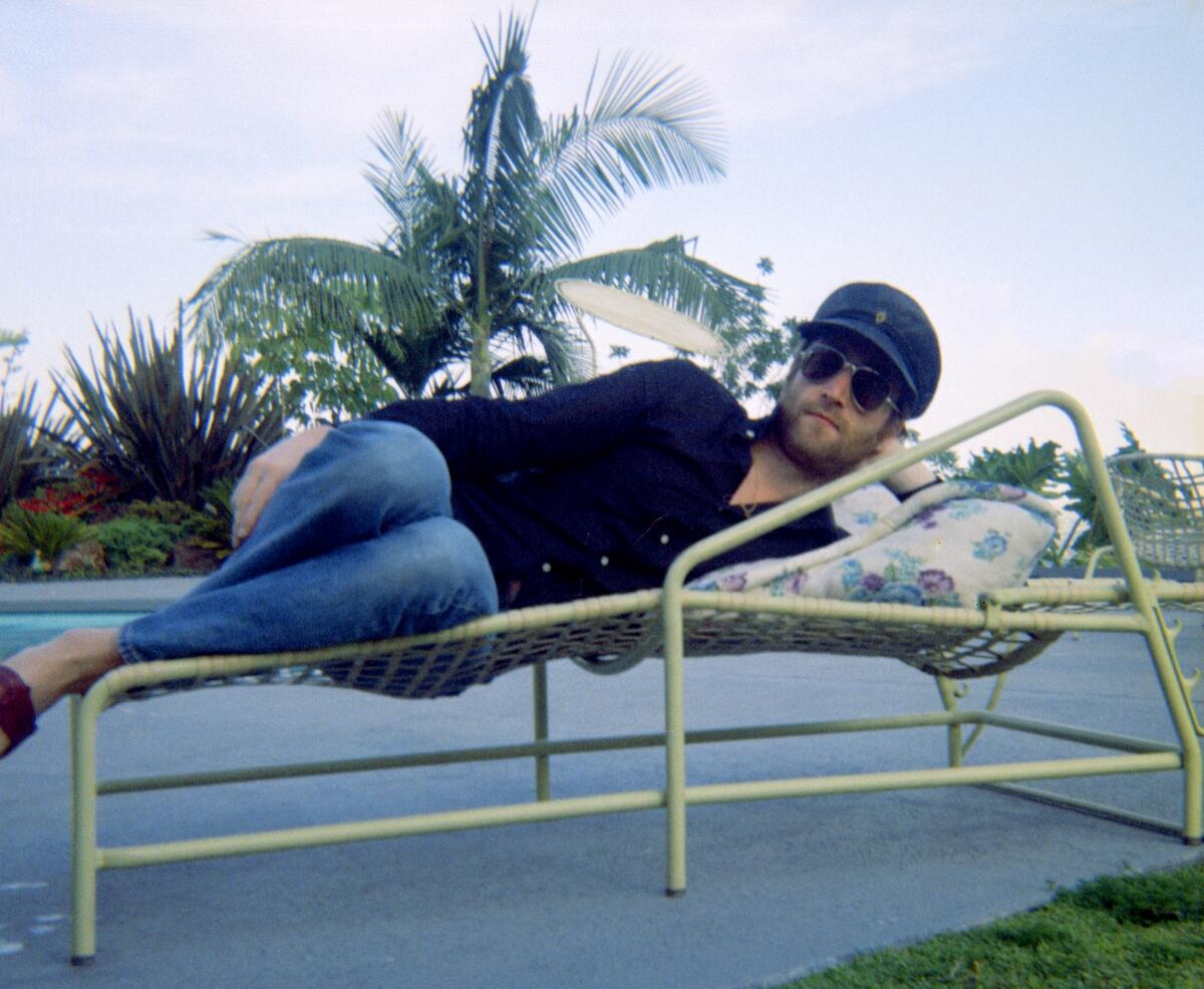 John Lennon relaxing on patio furniture by the swimming pool with palm trees in the background.