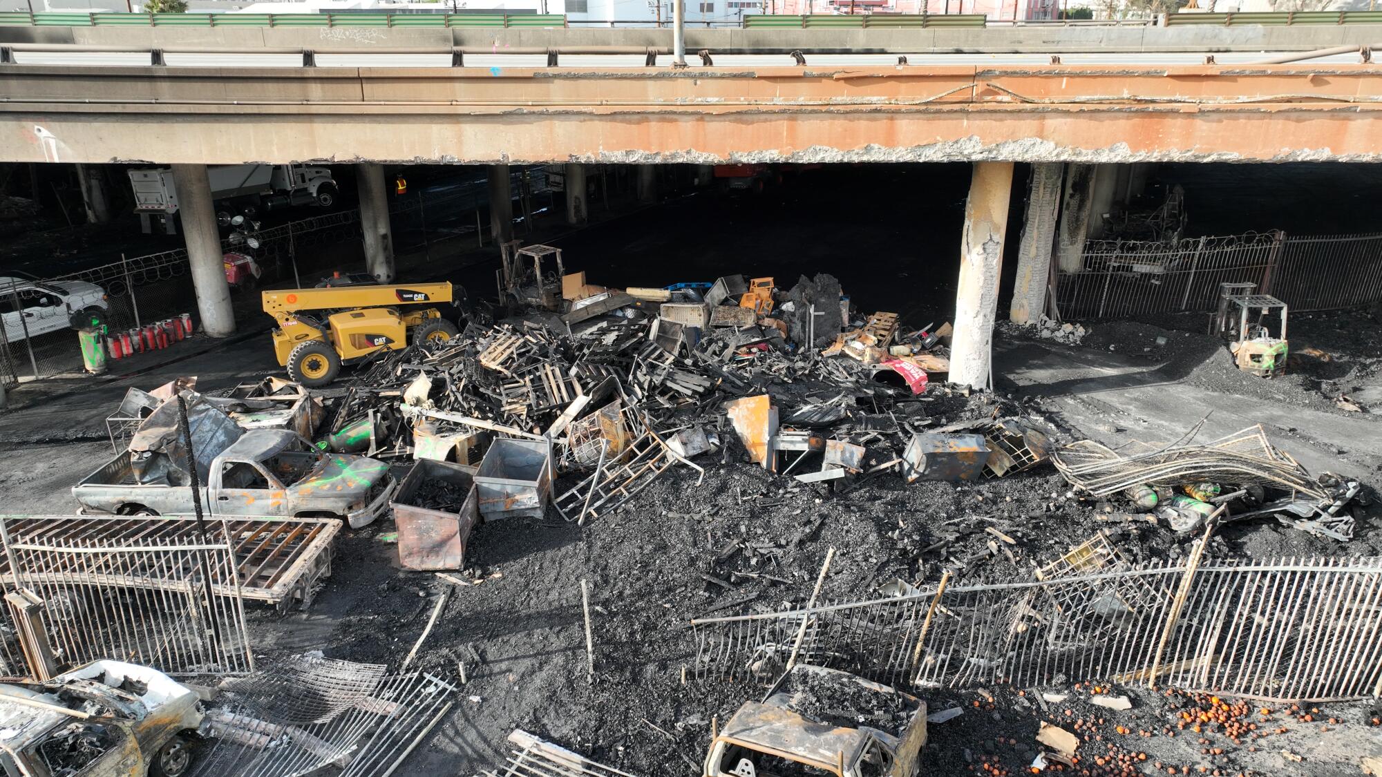 An aerial view of a large pile of charred debris between a stretch of freeway and what remains of a tall iron fence and gate