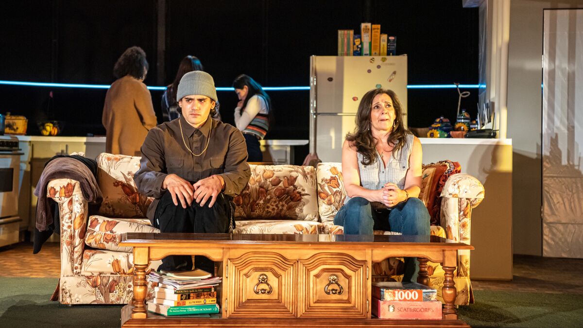 A young man in a knit cap and a crying woman sit side by side on a sofa in a play rehearsal.
