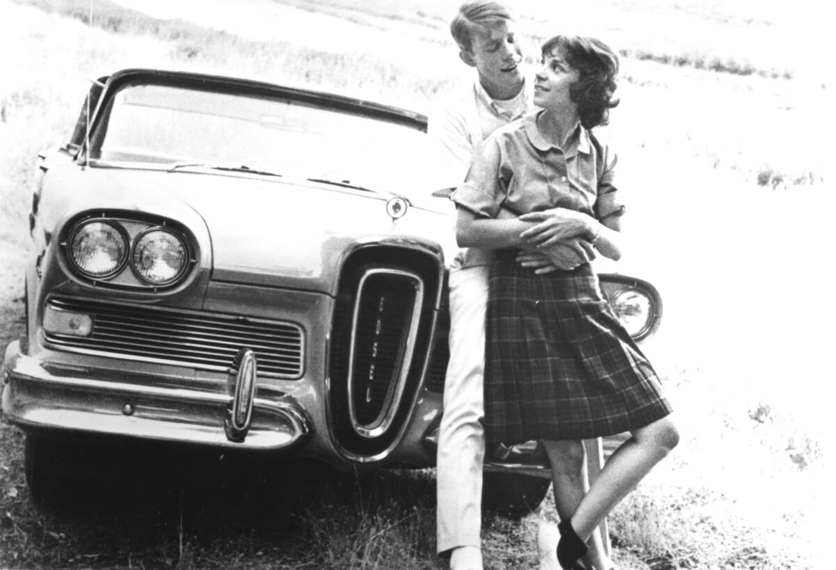A black-and-white photo of two young adults, a man with his arms around a girl's waist, leaning against a car.