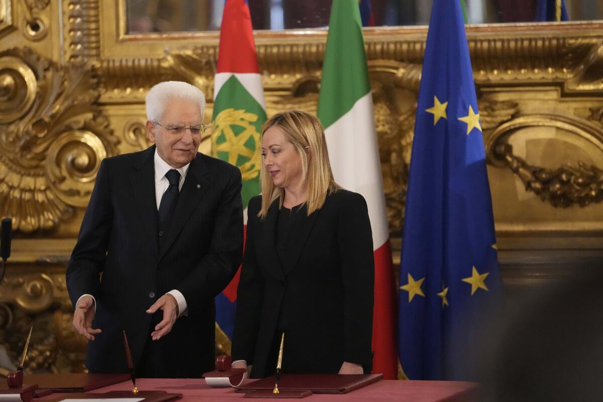 A man in a black suit stands next to a woman in a black suit as they stand in front of three multicolored flags.