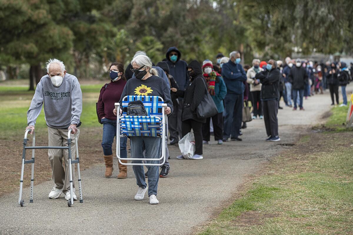 An elderly man using a walker at the front of a long line of people standing in a park