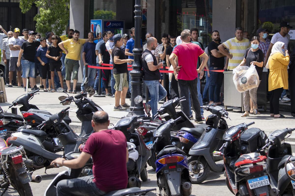 Behind parked motorbikes, people stand in a close-packed line, a few wearing masks.