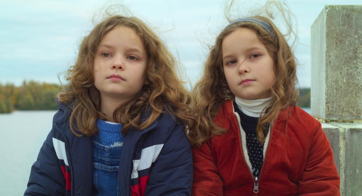 Two young girls who look alike sit outside with the water in the background
