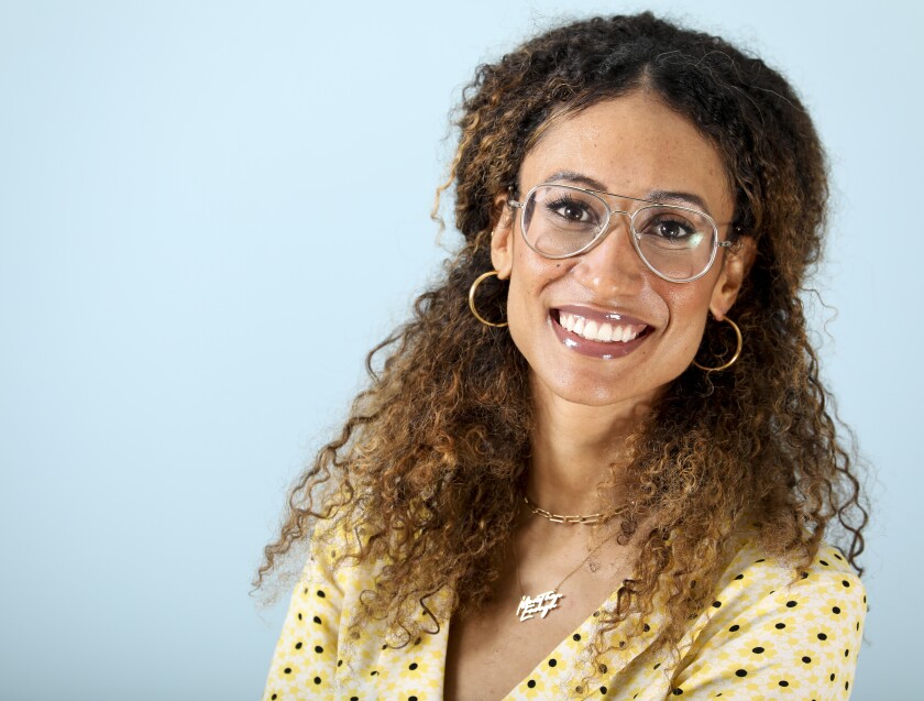A woman with curly brown hair and glasses in a yellow patterned shirt smiles.