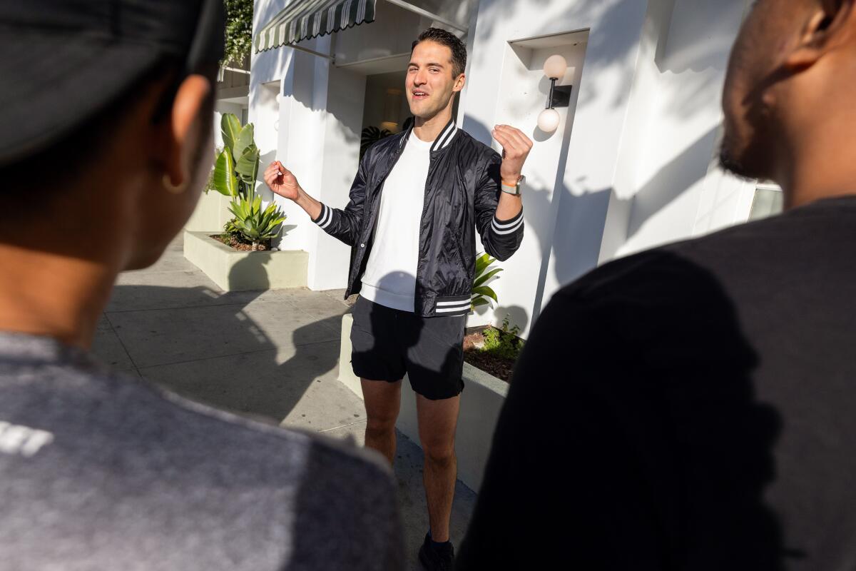 A man in shorts, T-shirt and open zip jacket stands outside a building gesturing enthusiastically.